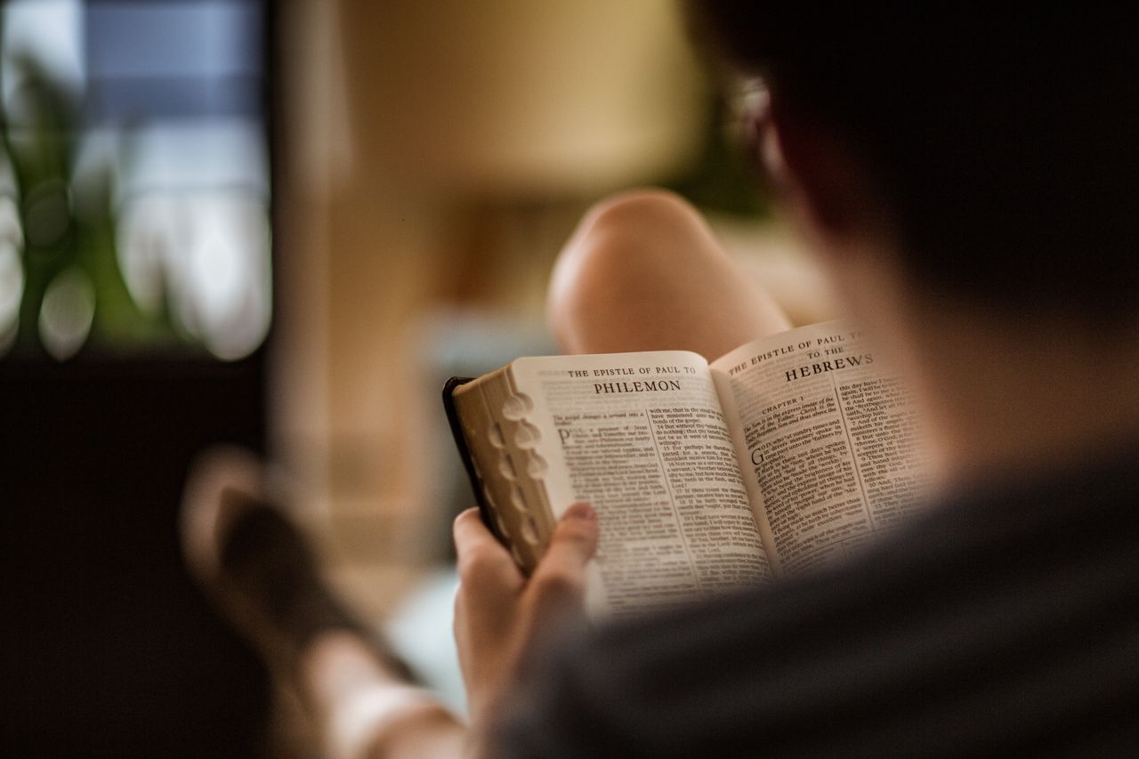 egy kanapén ülő férfi a Szent Bibliát tanulmányozza, amelynek az oldalai Pál Filemonhoz írt levelénél vannak nyitva